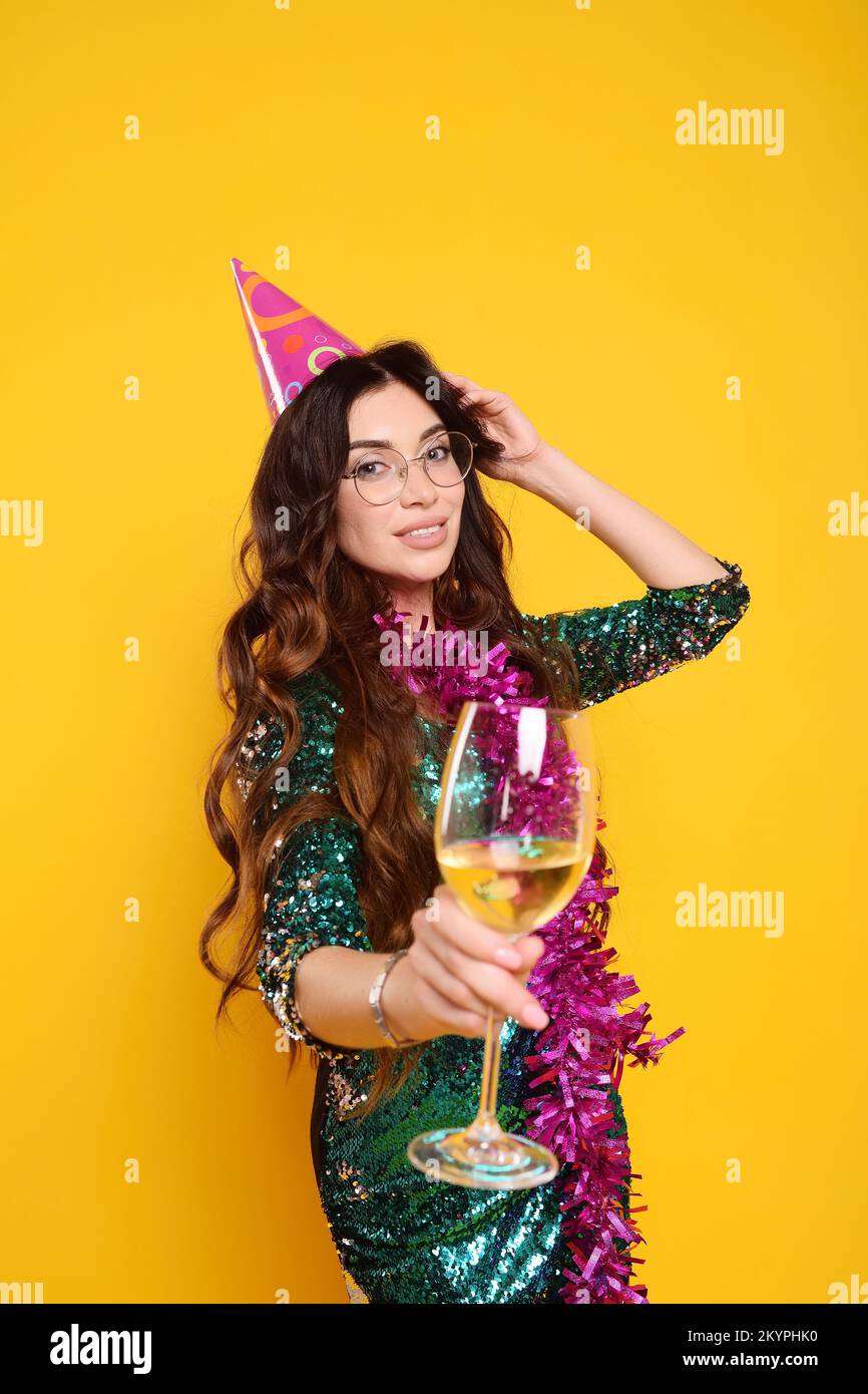 Eine Frau in einem eleganten Kleid in einer Partymütze mit einer pinkfarbenen Boa um den Hals und einem Glas Champagner, das auf einem gelben Hintergrund lächelt Stockfoto
