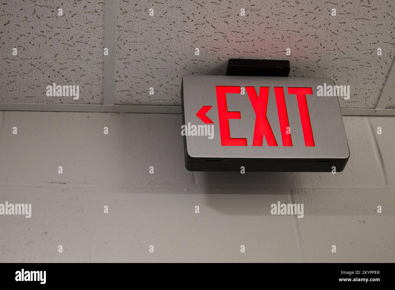 Ausgangsschild im US-Schulgebäude mit bemalter Wand und abfallender Decke. Die Wörter EXIT sind hellrot und der Richtungspfeil zeigt nach links. Stockfoto