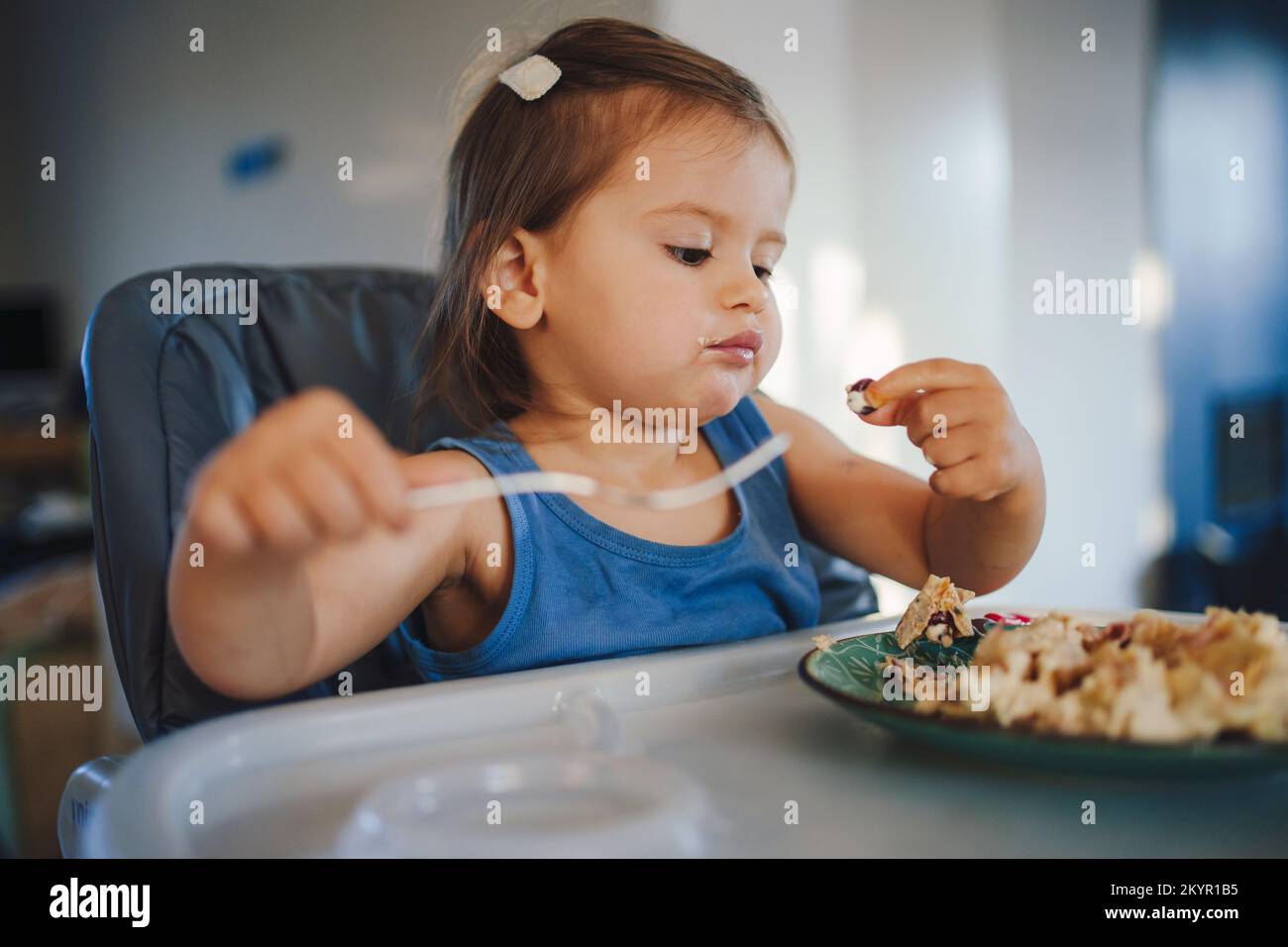 Ein glückliches kleines Mädchen lernt, sich selbst mit einer Gabel zu essen, während es im Kinderstuhl sitzt. Ein lächelndes Kind isst mit seinen Händen. Stockfoto