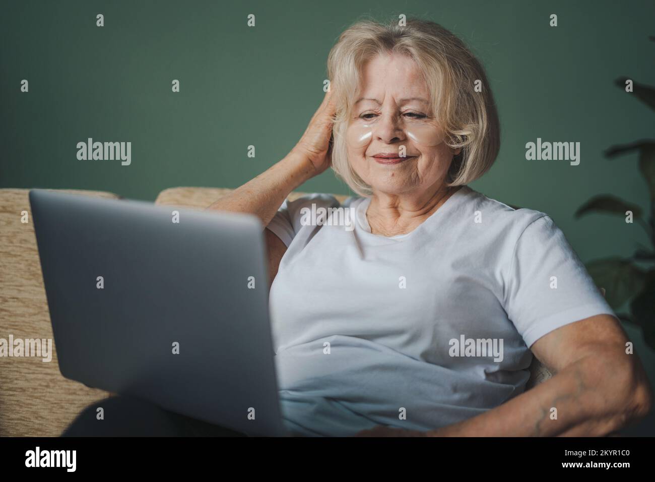 Fröhliche ältere, grauhaarige Rentnerin, die auf einem gemütlichen Sofa im Wohnzimmer sitzt und mit einem Laptop im Internet surft. Online-Kommunikation Stockfoto