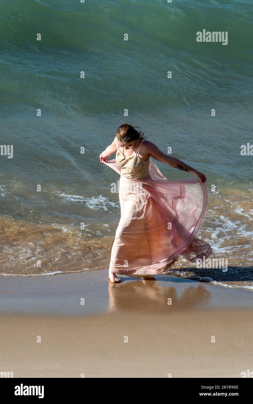 Wunderschöne junge Frau in fließendem Kleid, tanzt entlang der Wellen an einem nassen Sandstrand Stockfoto