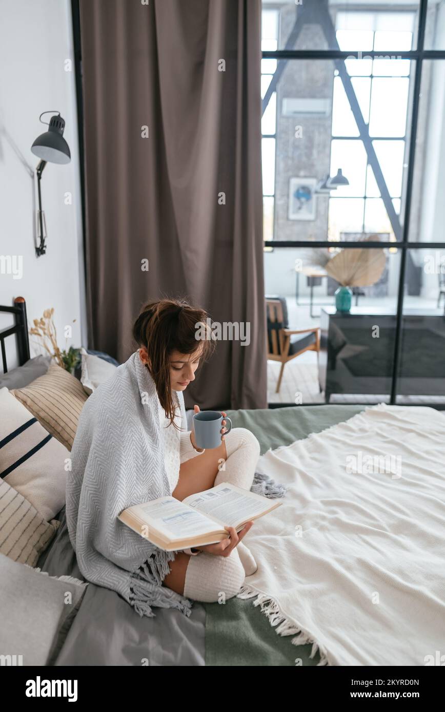 Frau liest Buch und genießt warmen Kaffee in einer sonnigen Wohnung. Stockfoto