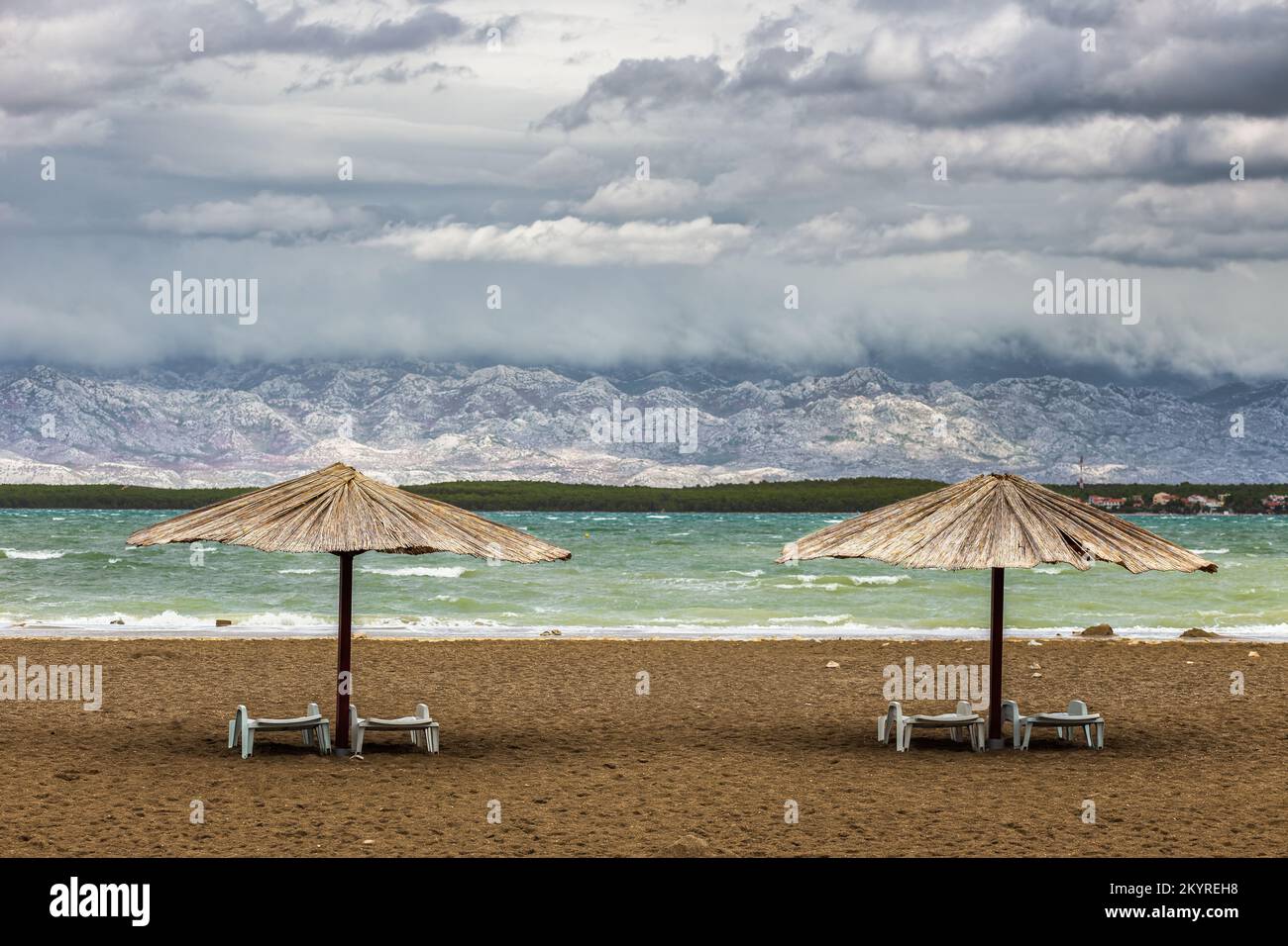 Nin, Kroatien - zwei Schilf-Sonnenschirme am leeren Queen's Beach in der mediterranen Stadt Nin am Ende des Sommers mit Velebit-Bergen im Hintergrund Stockfoto