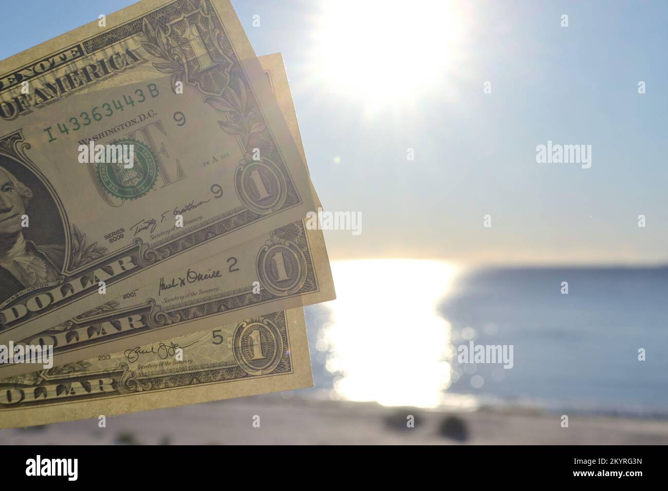 Person, die in der Hand Papierrechnungen von Dollar mit Nennwert von einem Dollar auf dem Hintergrund der hell leuchtenden Sonne, blauen Himmel und ruhiges blaues Meer hält. Ein-Dollar-Banknoten vor dem Hintergrund des blauen Meereshimmels Stockfoto