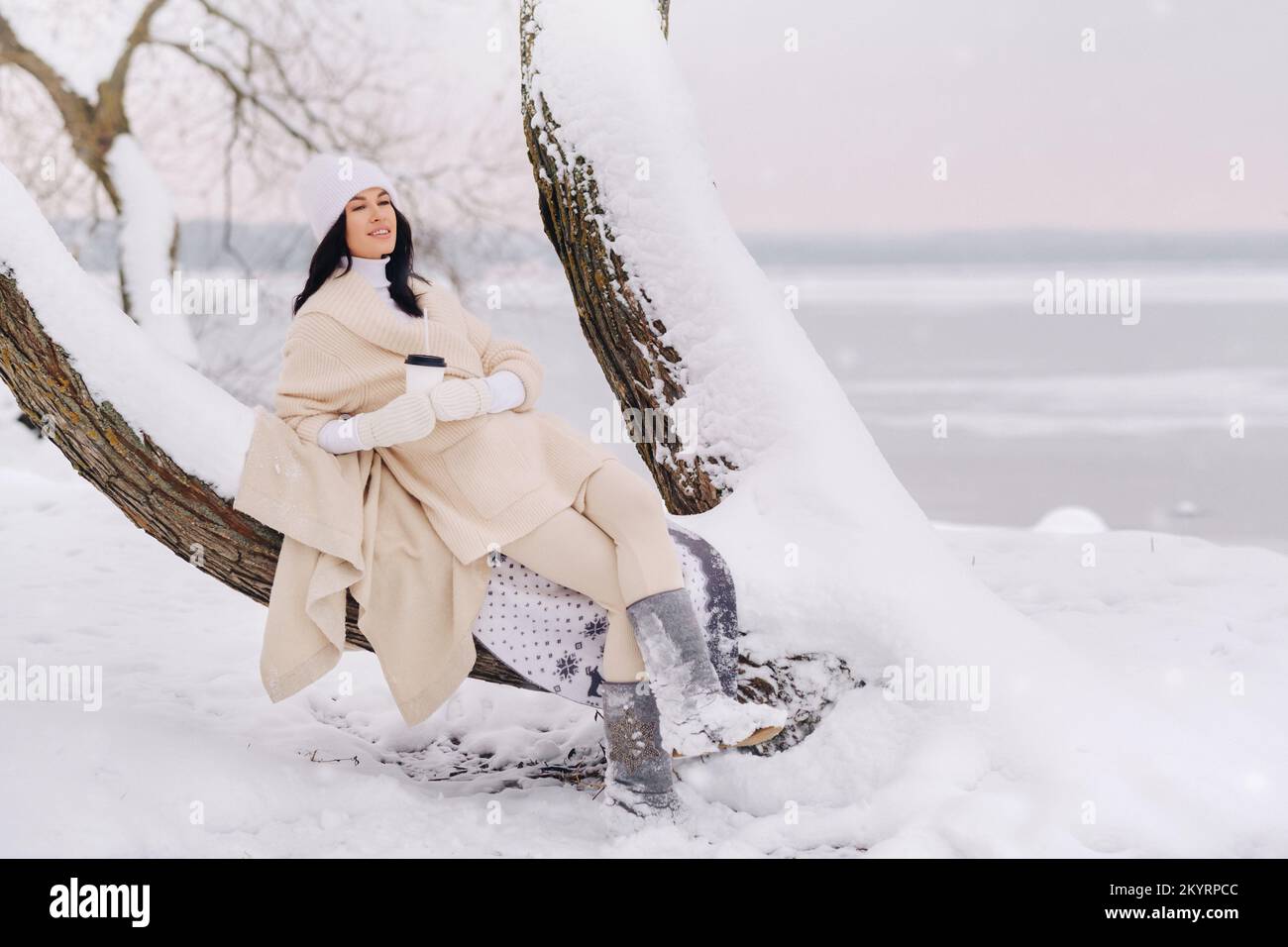 Ein hübsches Mädchen mit einer beigen Strickjacke und einem weißen Hut, das Tee in einem verschneiten Winterwald in der Nähe eines Sees trinkt Stockfoto