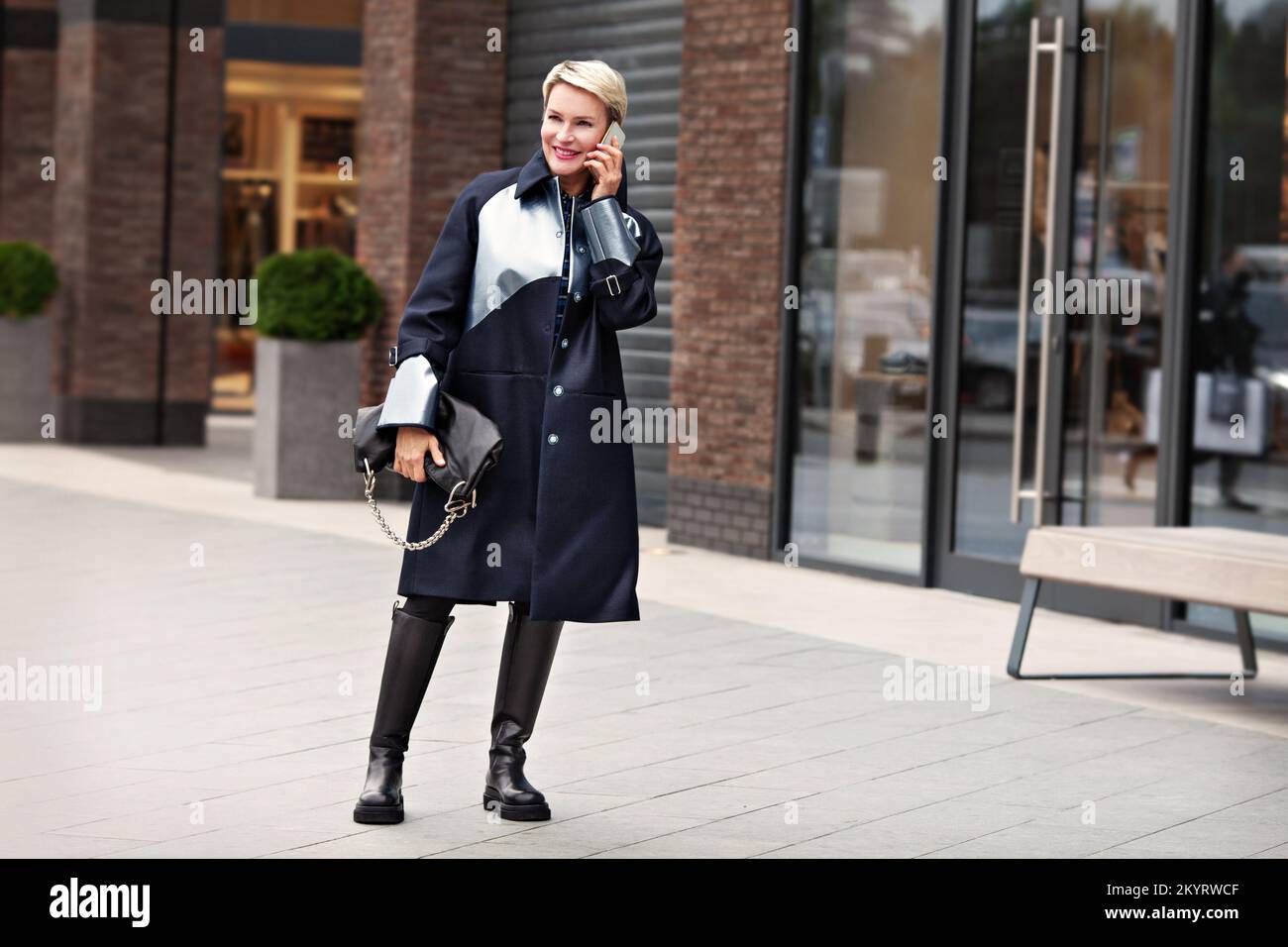 Modefrau in trendigen Outfits, schwarzer Mantel, Lederstiefel, Leggings. Das stilvolle, modische Modell spricht und lächelt auf der Straße in der Nähe Stockfoto