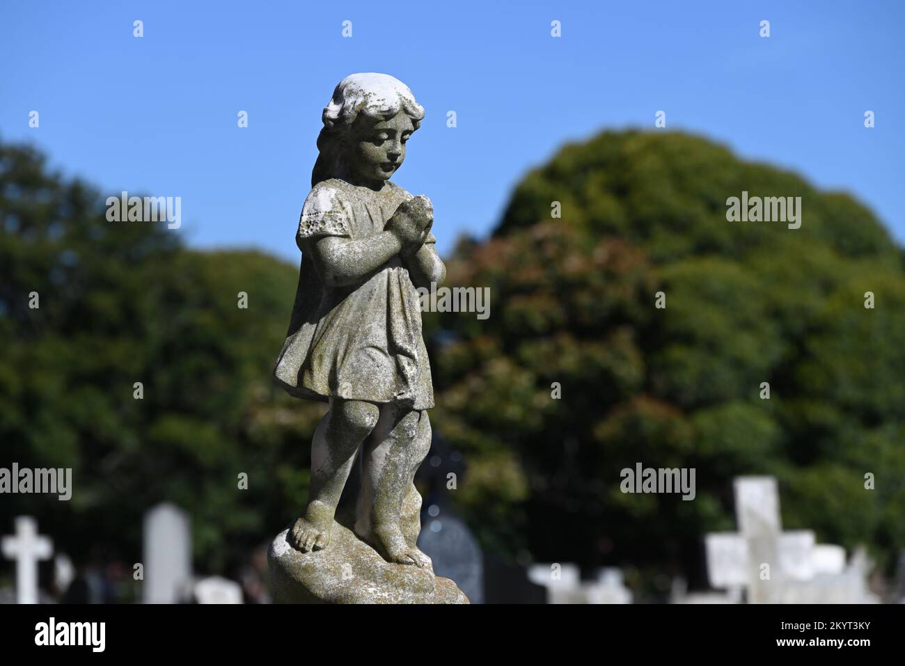 Alte, abgenutzte und verwitterte Steinskulptur eines umhüllten Kindes, das trauernd nach unten schaute, Hände zusammengefesselt im Gebet, auf einem Friedhof Stockfoto