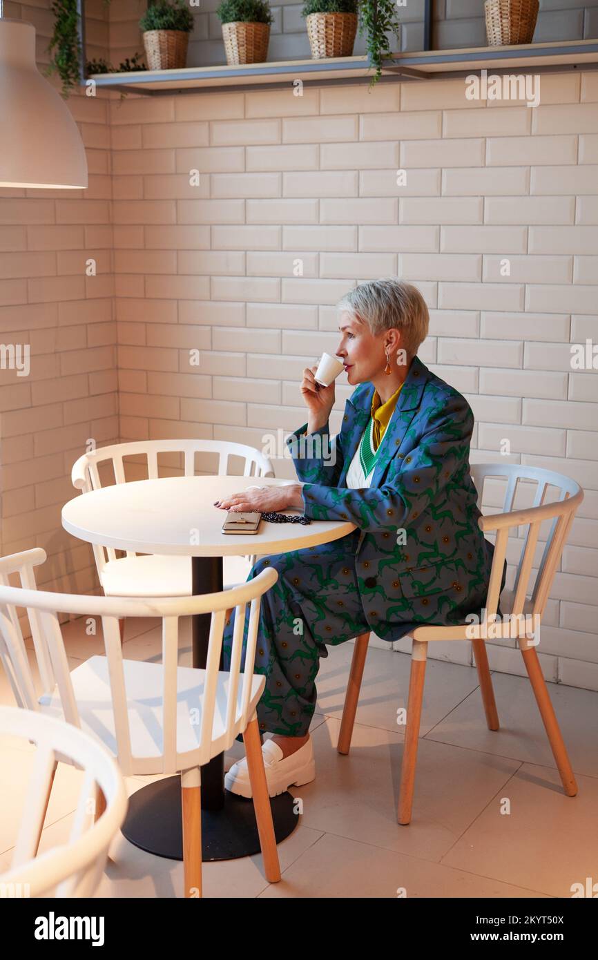 Reife Geschäftsfrau, die Espresso trinkt und in einem hellen Café nachdenkt. Stilvolle Frau in leuchtend bedruckten Kostümen und kurzen grauen Haaren Stockfoto