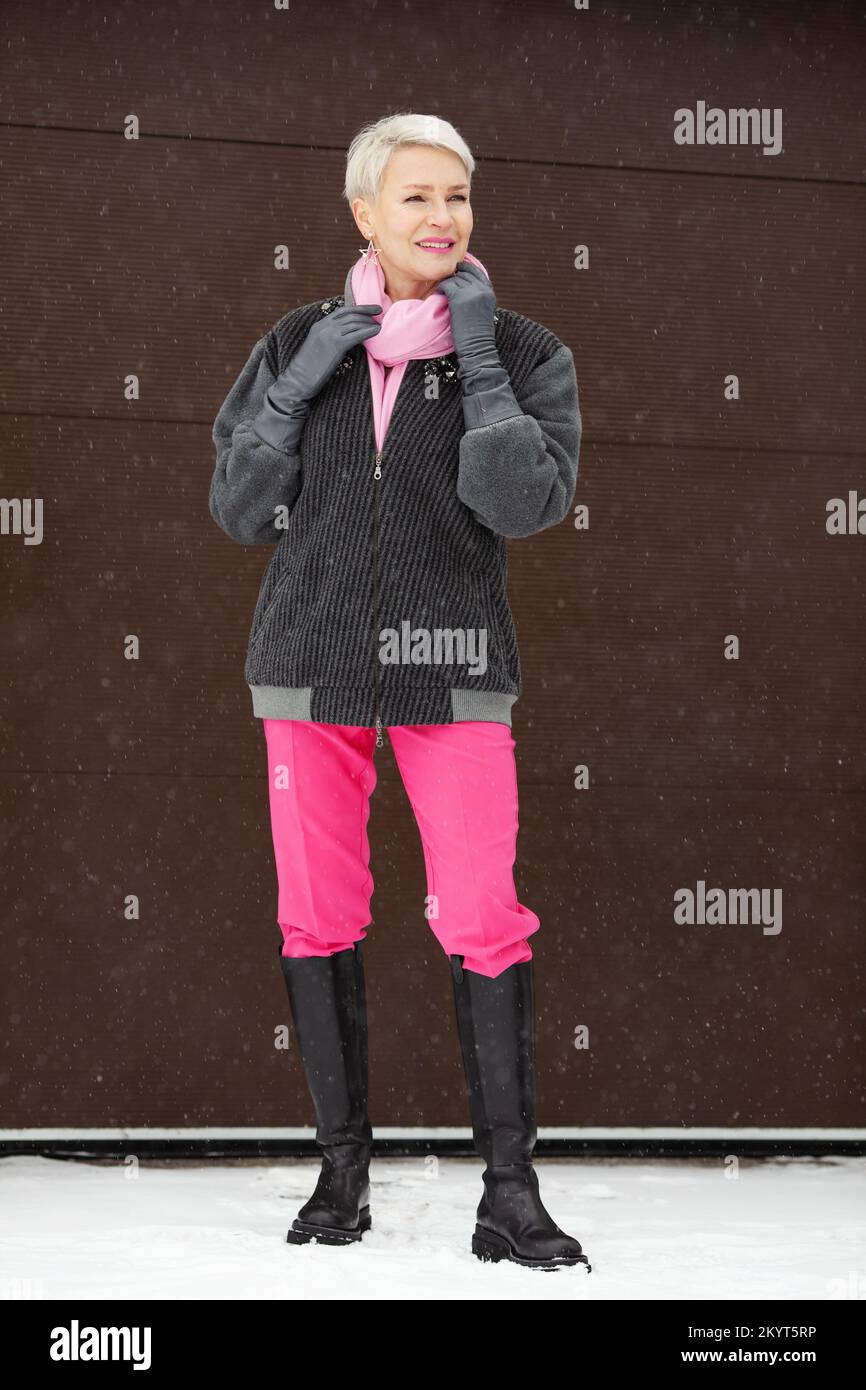 Winteroutfit für ältere Frauen mit hohen Stiefeln, pinkfarbenen Hosen, Bomberjacke, stilvollem, durchgehendem Look einer älteren Frau mit kurzen Haaren und leuchtenden Details Stockfoto