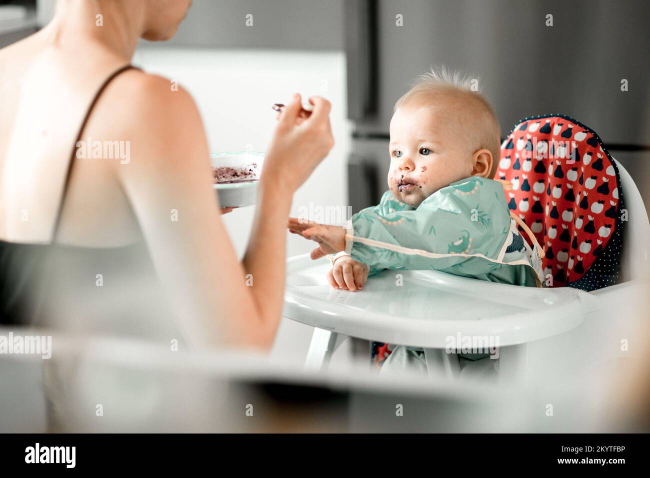 Mutter Löffel Fütterung ihres kleinen Jungen Kind in Babystuhl mit Fruchtpüree in der Küche zu Hause. Baby feste Nahrung Einführung Konzept Stockfoto