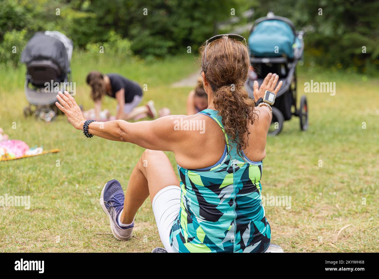 Selektive Fokussierung vom Rücken einer braunen Trainerin, die Yoga-Posen übt und trainiert, während sie auf dem Gras sitzt und von Schülern in der Natur umgeben ist Stockfoto