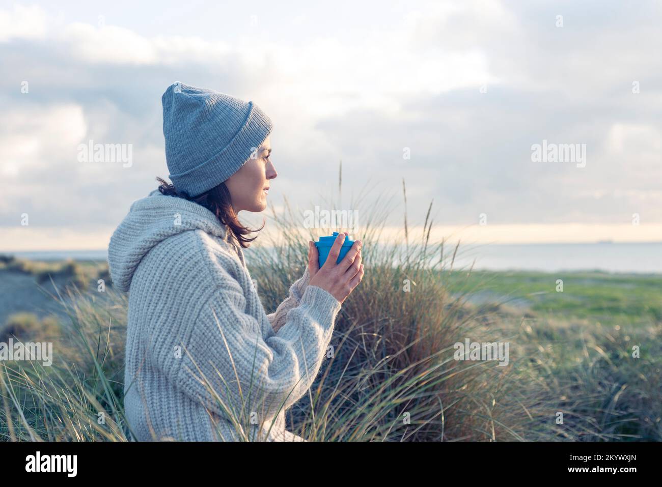 Eine Frau in Strickmode, die ein heißes Getränk genießt und dabei auf das Meer blickt. Stockfoto