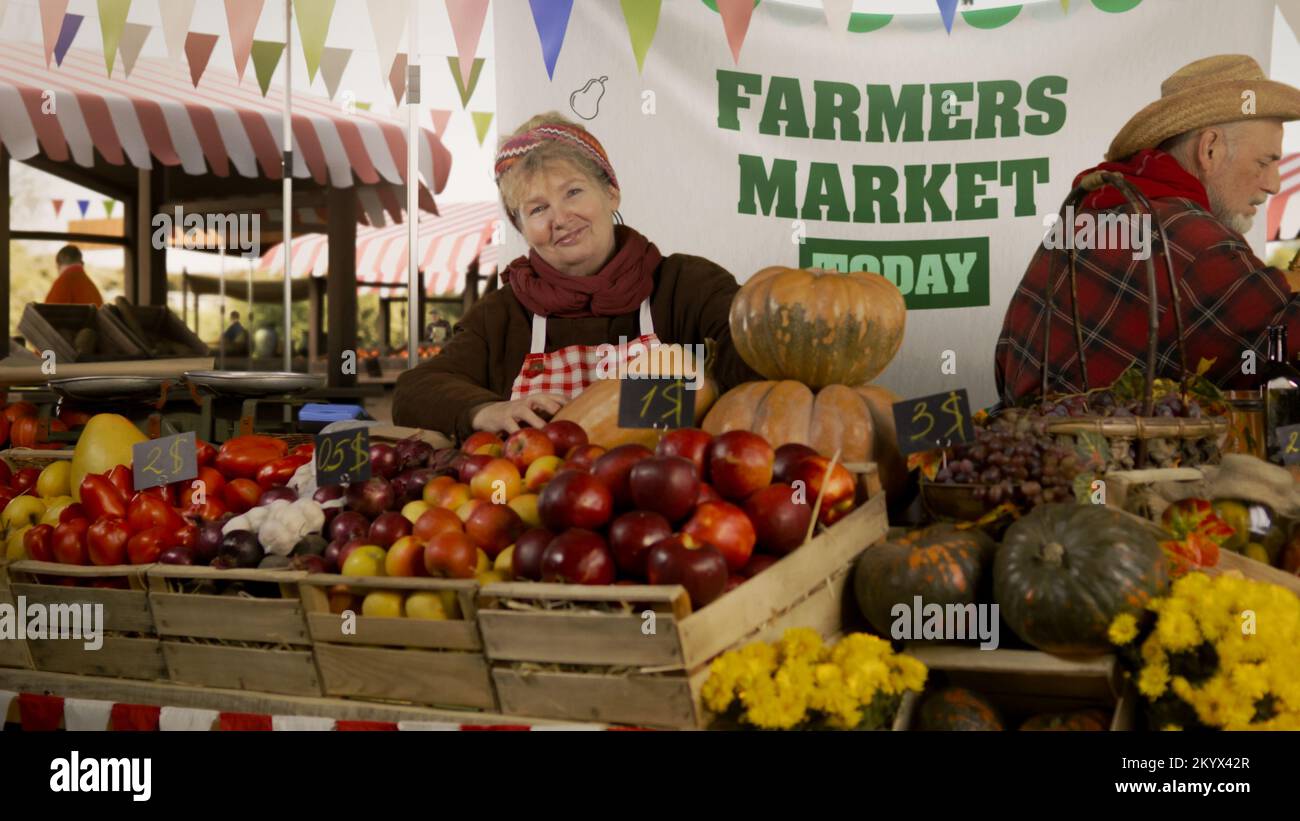 Die Bauernfrau steht am Stand mit frischem, bunten Obst und Gemüse, schaut auf die Kamera. Farmers Market Festival. Vegetarische, biologische und gesunde Lebensmittel. Landwirtschaft. Stockfoto