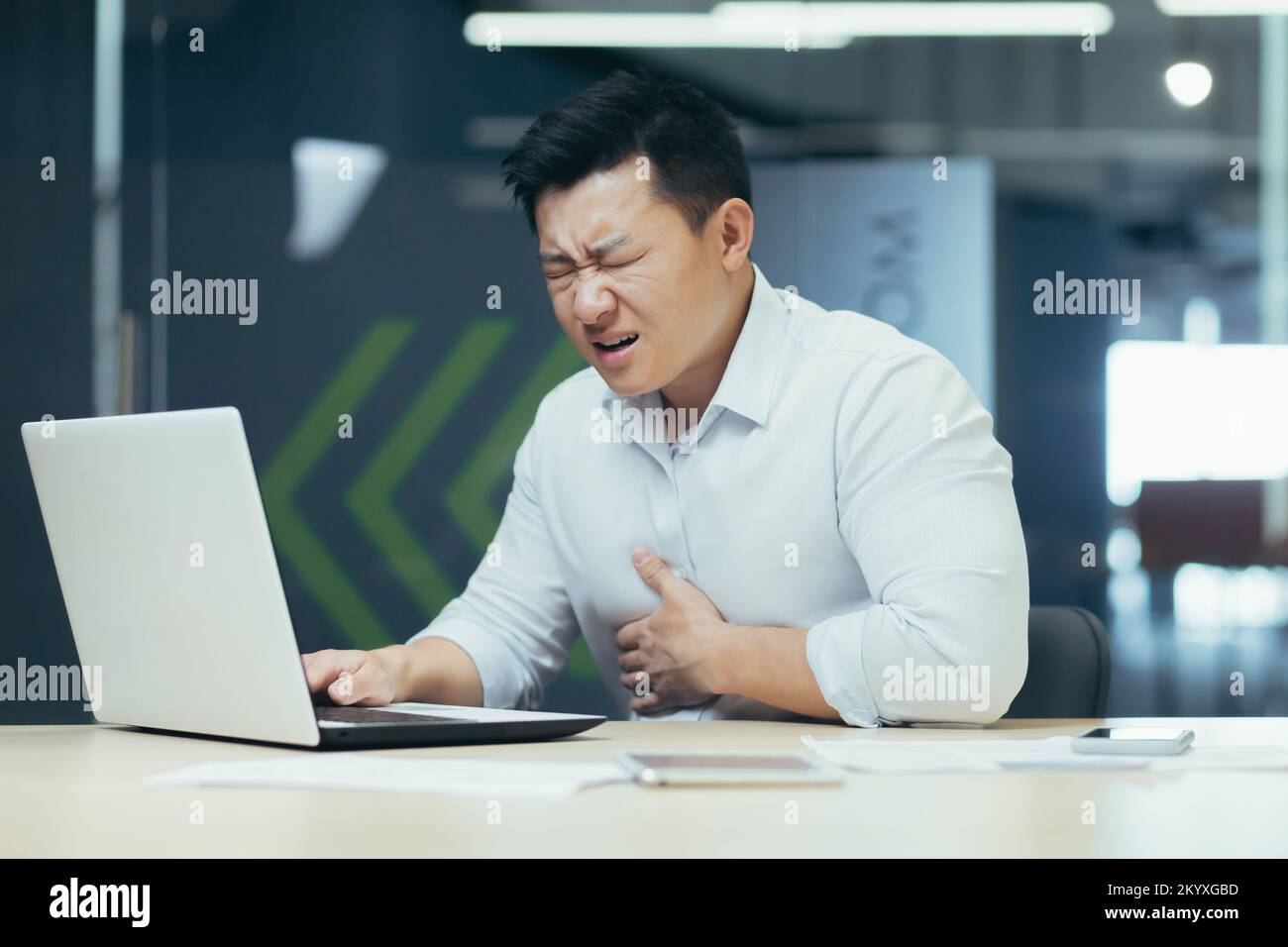 Herzinfarkt am Arbeitsplatz. Ein junger asiatischer Mann sitzt im Büro am Tisch, hält seine Brust, grimmig, fühlt Schmerzen, hat einen Schlaganfall. Stockfoto