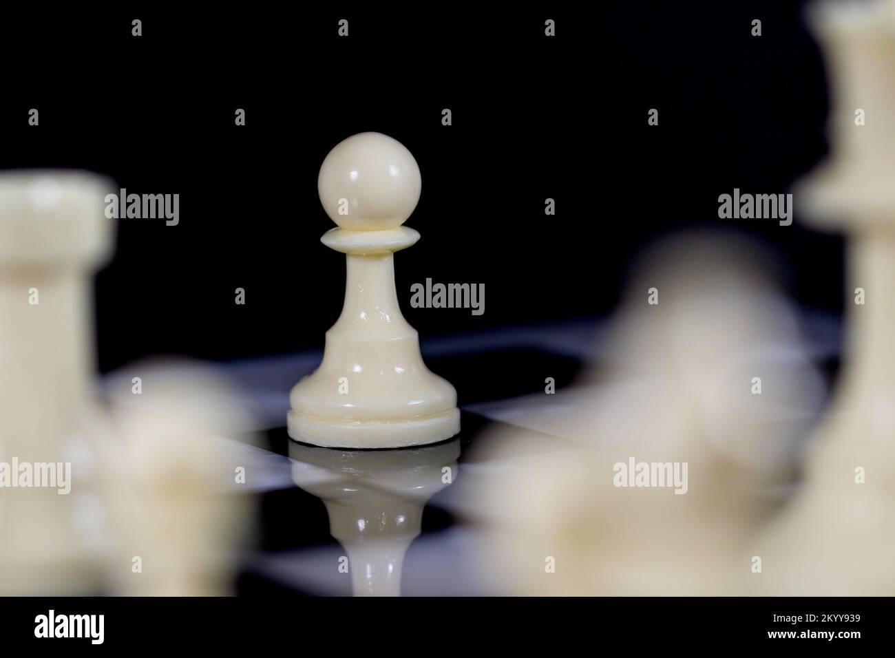 Das kleinste davon: Ein einsamer weißer Bauer steht hinter anderen weißen Schachfiguren; Reflexion in einem schwarz-weißen, marmornen Schachbrett. Schwarzer Hintergrund Stockfoto