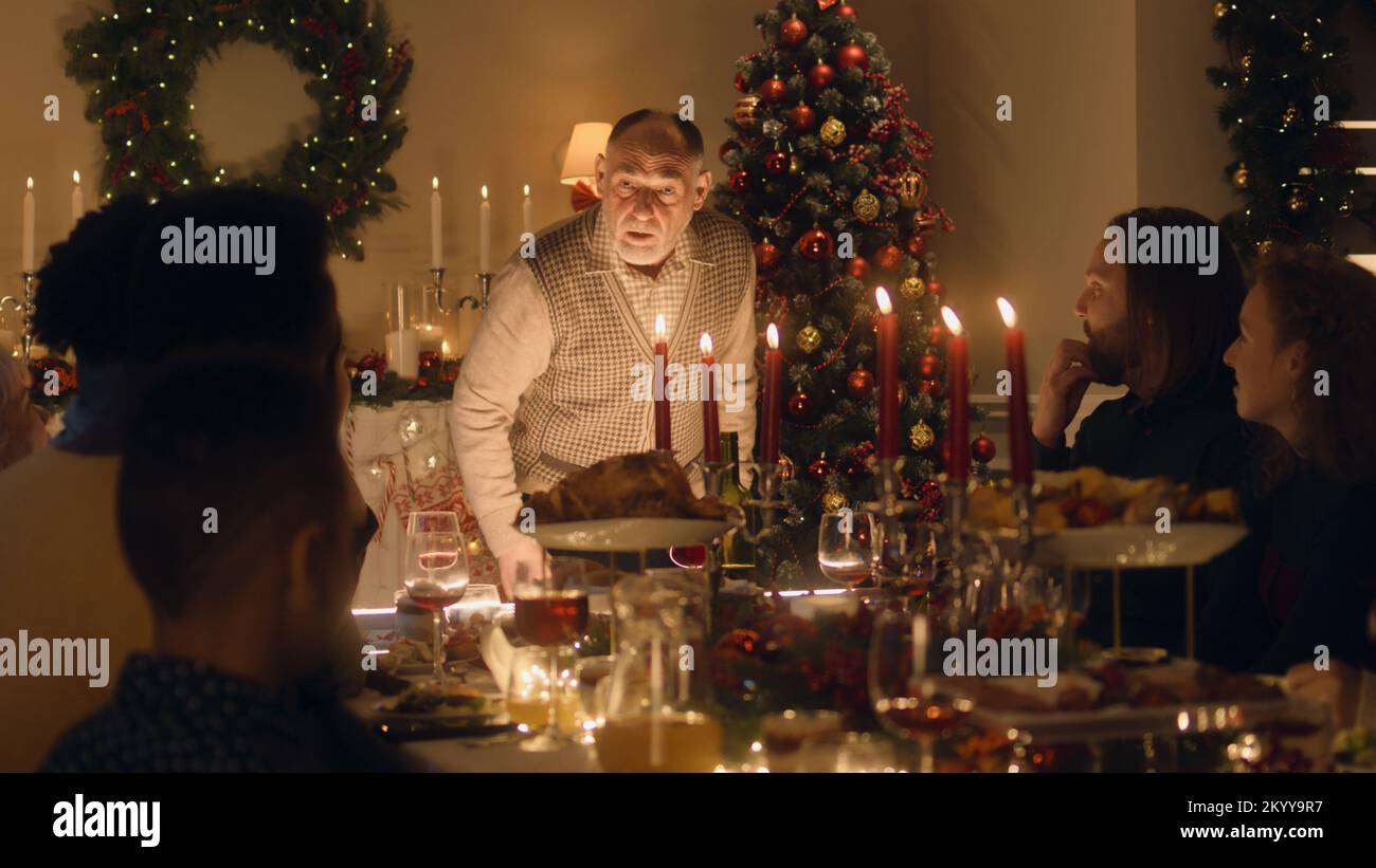 Älterer Großvater erzählt seiner großen multikulturellen Familie Witze oder eine Lebensgeschichte. Sie feiern Weihnachten. Serviert wird ein Tisch mit Geschirr und Kerzen. Warme Atmosphäre beim Weihnachtsessen mit der Familie zu Hause. Stockfoto