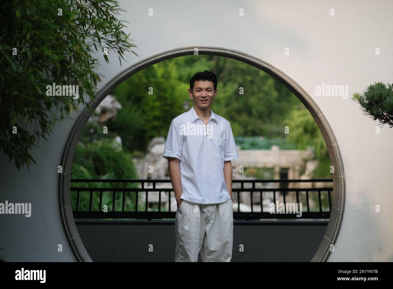 Süßer, lächelnder chinesischer junger Mann am Mondtor im traditionellen chinesischen Garten. Vorderansicht Stockfoto