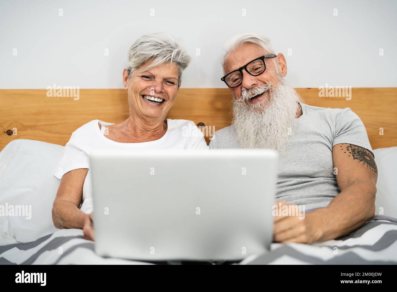 Glückliches Seniorenpaar, das Spaß mit dem Laptop hat, während es im Bett unter Decken liegt - Technologie und Lifestyle-Konzept für ältere Menschen Stockfoto