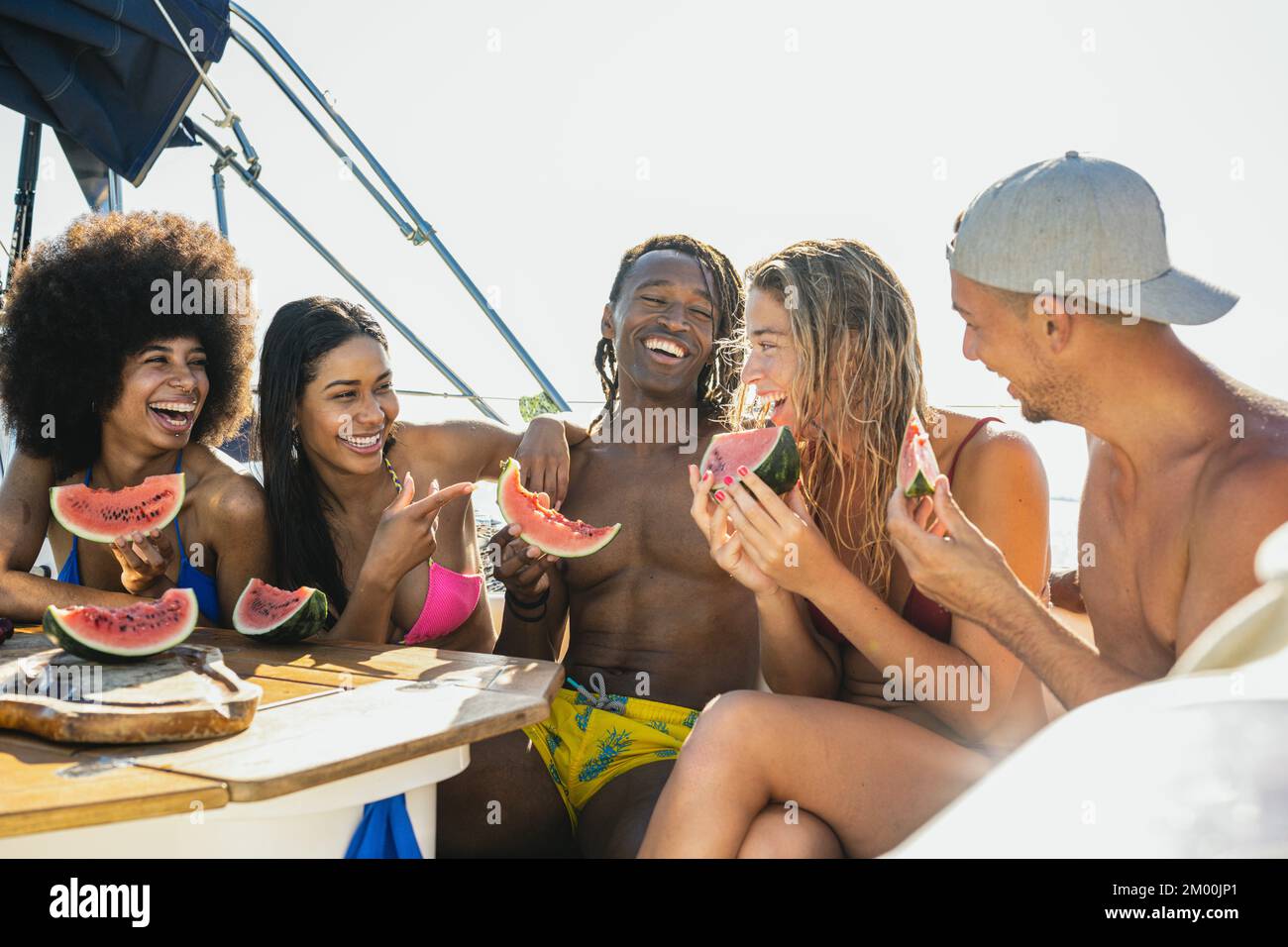 Multirassische Gruppe von Menschen, die Spaß auf dem Yachtdeck haben und lachen, fröhliche Männer und Frauen, die Obst essen und auf einer Sommerparty auf dem Boot lächeln Stockfoto