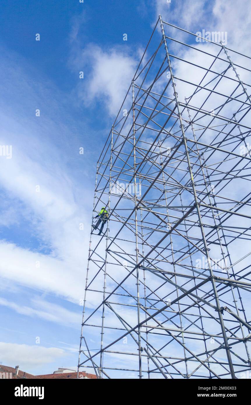 Unidentifizierter Arbeiter in der Höhe, der einen Gerüstturm demontiert. Blauer wolkiger Himmel im Hintergrund. Stockfoto