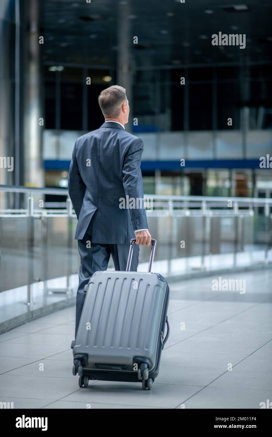 Geschäftsreise. Ein Mann im dunklen Anzug geht selbstbewusst mit dem Koffer, mit dem Rücken zur Kamera im Terminal. Stockfoto