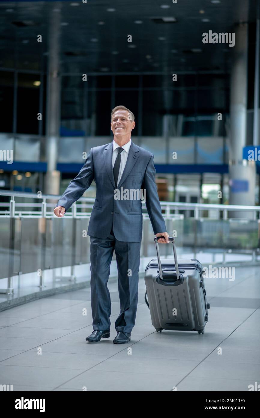 Trip, Aufregung. Lächelnder erfolgreicher Mann in elegantem dunklen Anzug, mit Trolley-Koffer, der aufgeregt im Terminal läuft. Stockfoto