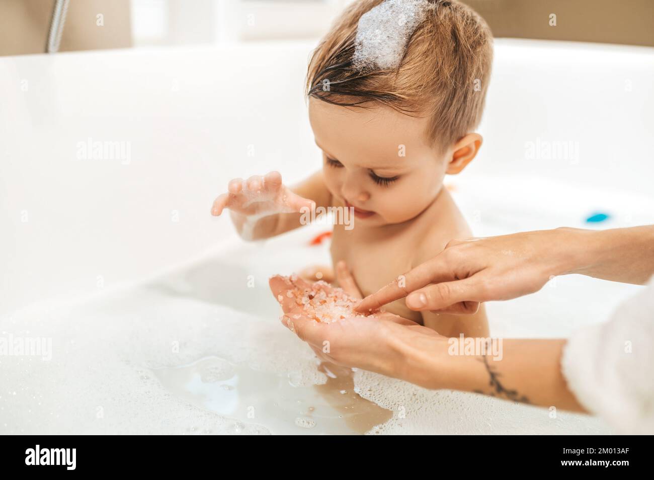Konzentriertes Baby sitzt im Seifenwasser und starrt auf die Kochkristalle der weiblichen Hand. Stockfoto