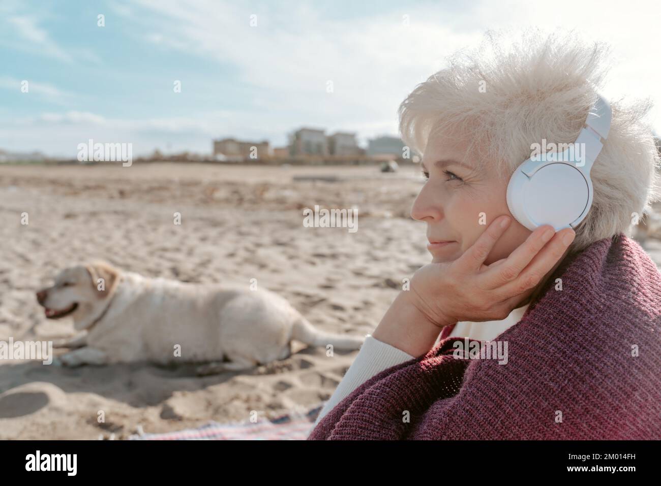 Lächelnde, grauhaarige Frau mit kabellosen Kopfhörern, die neben ihrem Haustier auf dem Sand saß. Stockfoto