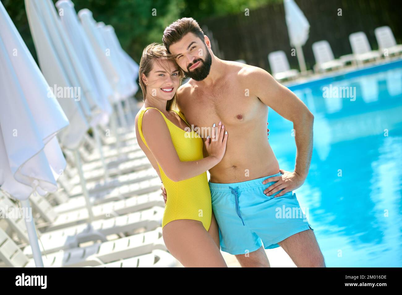 Eine lächelnde, wunderschöne Frau und ihr männlicher Begleiter stehen am Pool und posieren für die Kamera. Stockfoto