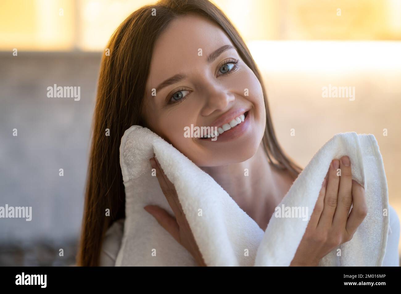 Zärtlichkeit. Nahaufnahme einer jungen, hübschen Frau mit einem zahnigen Lächeln, die auf die Kamera schaut und ihr Gesicht mit einem Handtuch im Haus berührt. Stockfoto