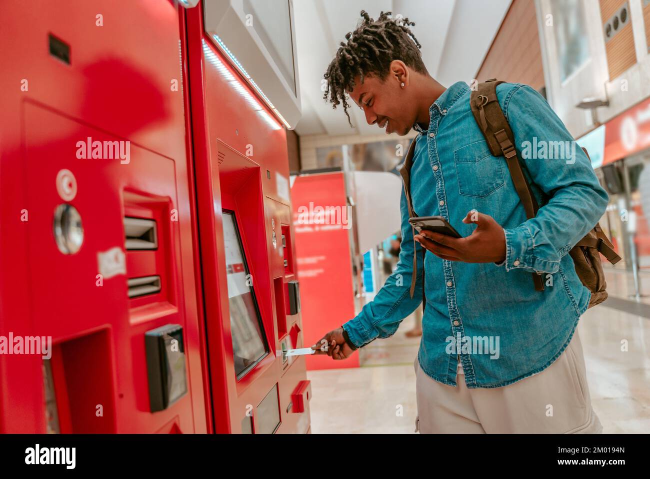 Kartenzahlung. Lächelnder afroamerikanischer Typ mit Smartphone und Rucksack, der die Karte in das Selbstbedienungs-Terminal am Bahnhof einsteckt. Stockfoto