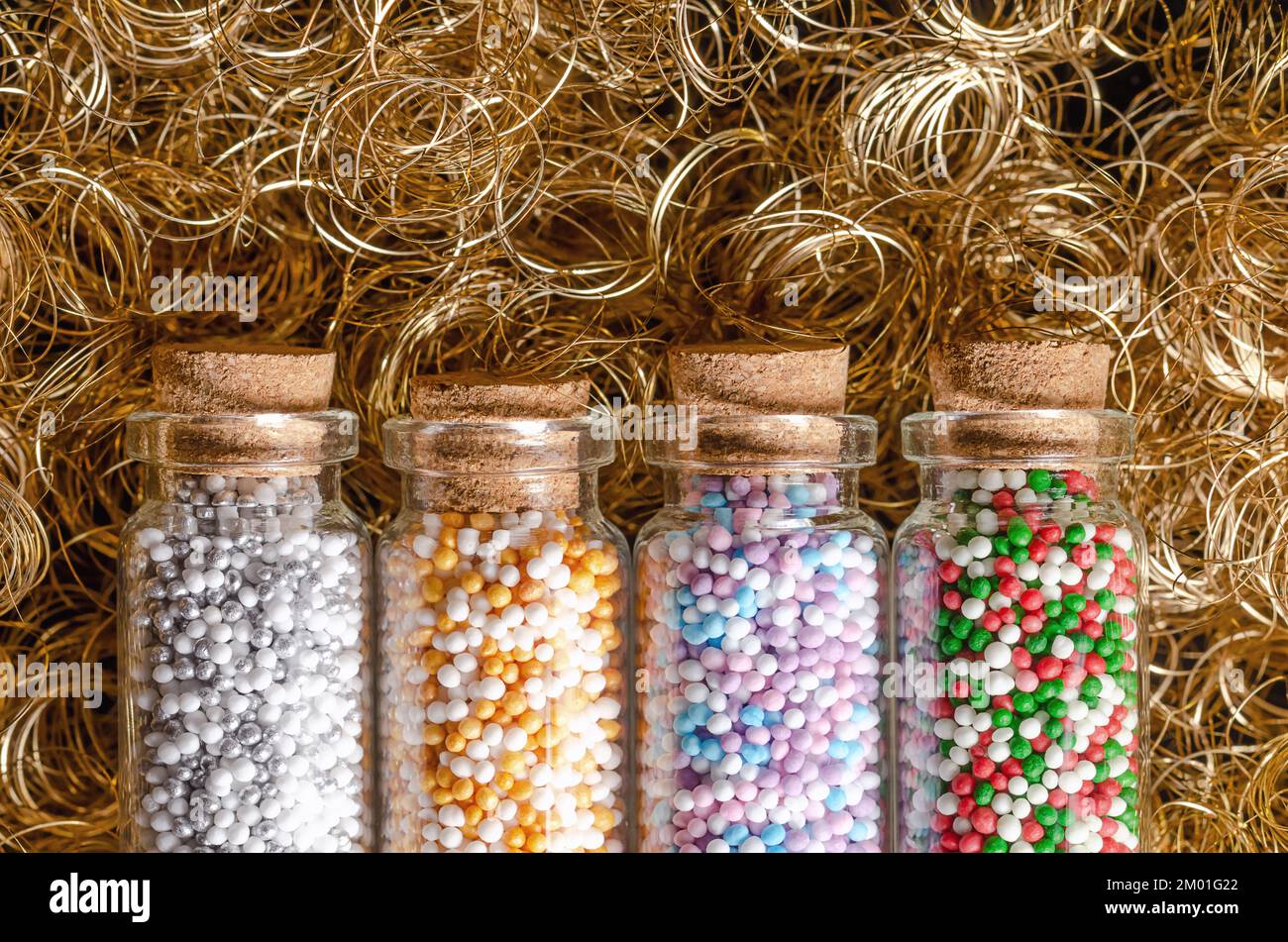 Nonpareils in kleinen Glasflaschen, über goldenem Engelhaar. Winter- und weihnachtsfarbene Mischungen von essbaren Süßwaren aus winzigen Zuckerbällen. Stockfoto