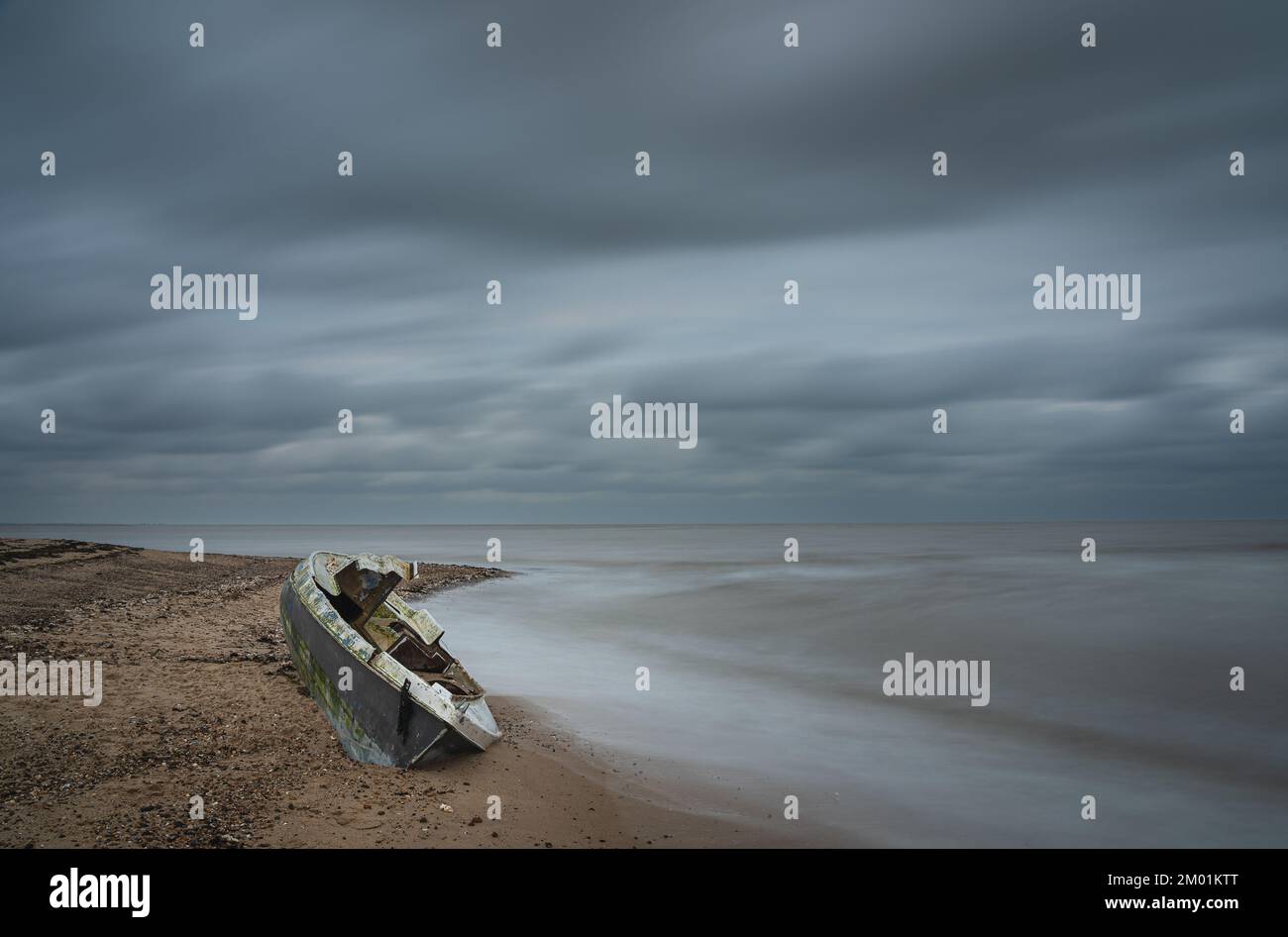 Altes, beschädigtes Boot am Strand auf Mersea Island in Essex. Langzeitaufnahme mit stimmungsvollem, bewölktem Himmel. Stockfoto