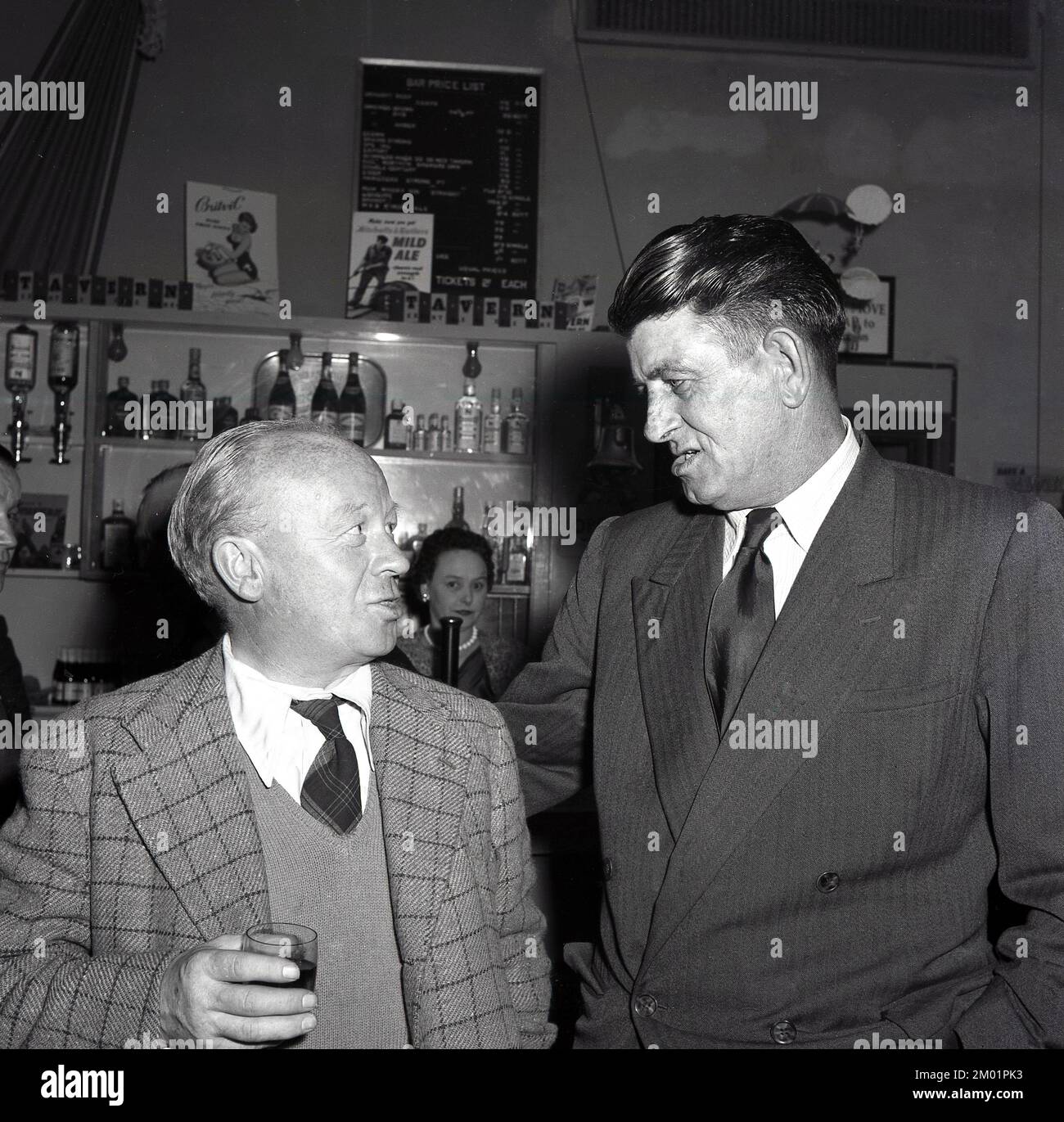 1950er, historisch, das ist ein Witz... nach der Arbeit und zwei Stahlarbeiter mit Hemd und Krawatte, die an einer gesellschaftlichen Veranstaltung teilnehmen und zusammen einen Drink in der Bar Port Talbot, South Wales, Großbritannien, trinken. Hinter der Bar ist eine Werbung für Mitchells & Butlers Mild Ale zu sehen. Stockfoto