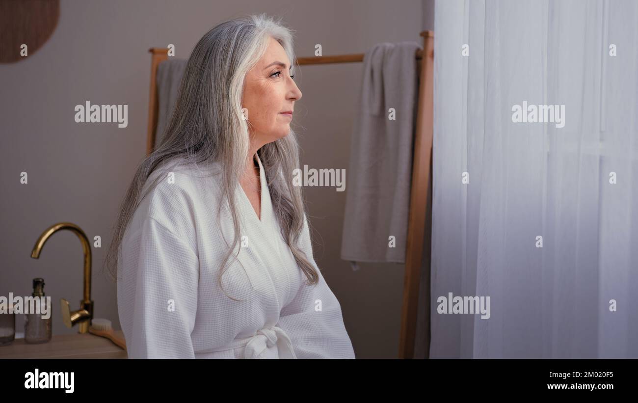Pensive ältere Dame, 60s alt, weiß, 50s ältere, reife, wunderschöne Frau im weißen Bademantel, die zu Hause im Badezimmer steht. Eine ruhige Großmutter Stockfoto
