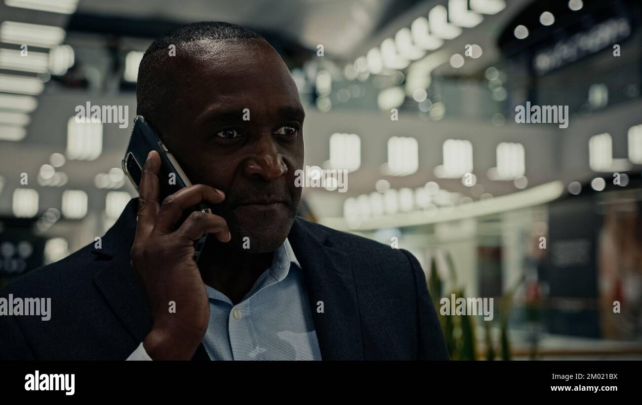 Laufender erwachsener Geschäftsmann, der mit seinem Mobiltelefon spricht, virtuelles Gespräch mit einem afroamerikanischen Geschäftspartner mittleren Alters, einem Angestellten Unternehmer im Einkaufszentrum Stockfoto