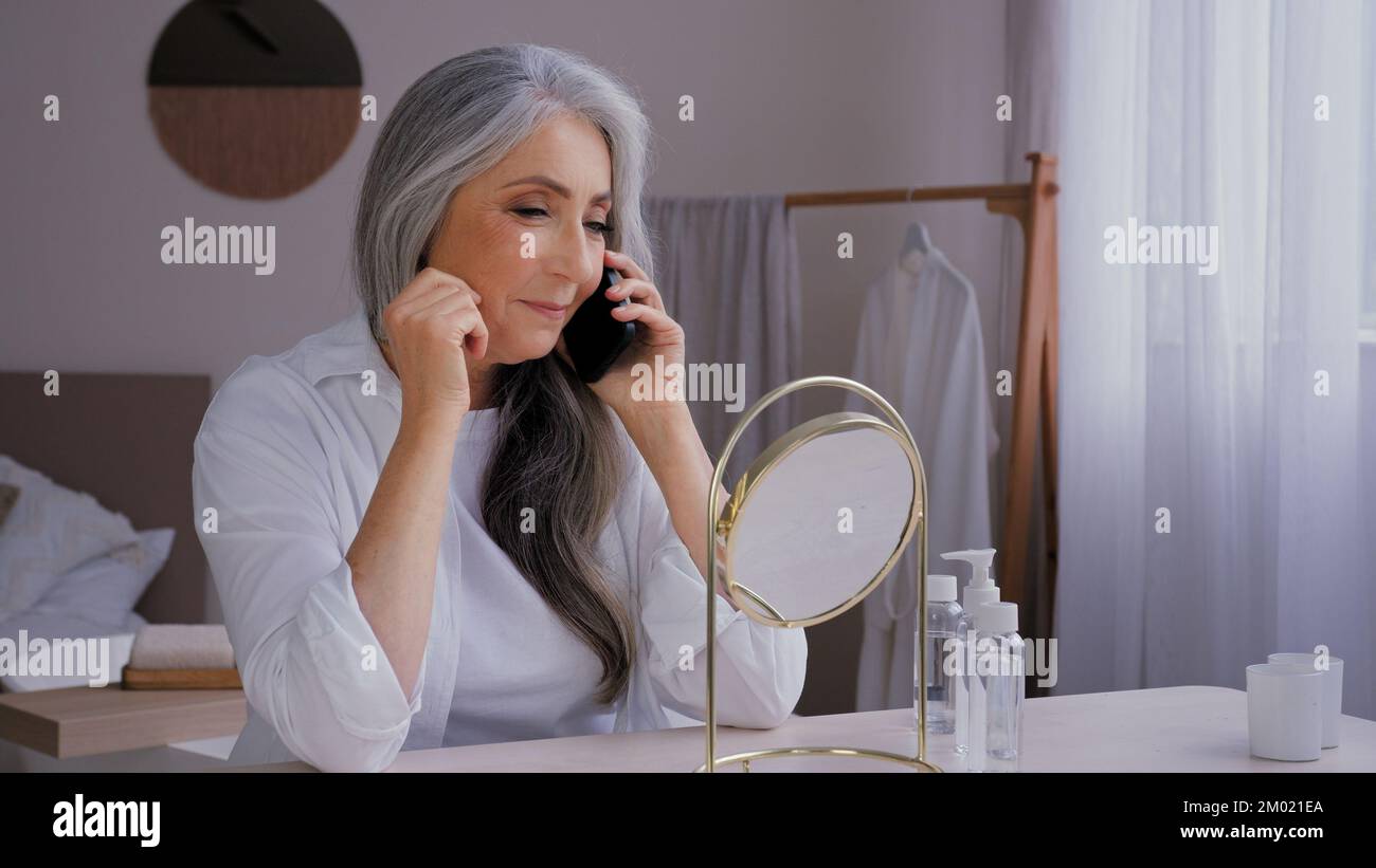 Ältere, grauhaarige, alte 60s-jährige Frau, die mit dem Handy spricht, Gesprächsgespräch im Spiegel, die Gesichtscreme mit dem Smartphone kauft Stockfoto