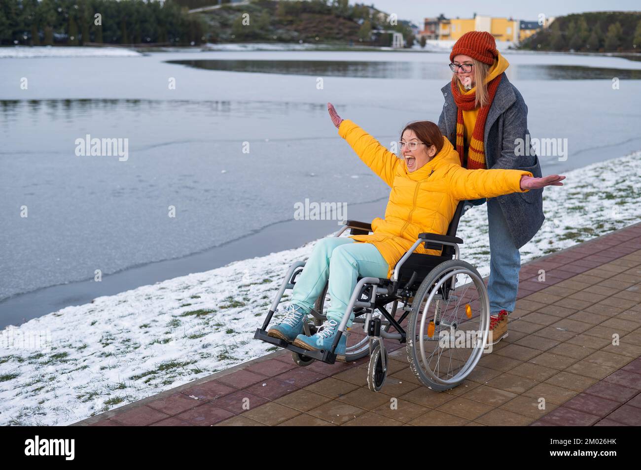 Kaukasische Frau schiebt ihre Freundin auf einen Rollstuhl am See. Frau fühlt sich wie Fliegen. Stockfoto