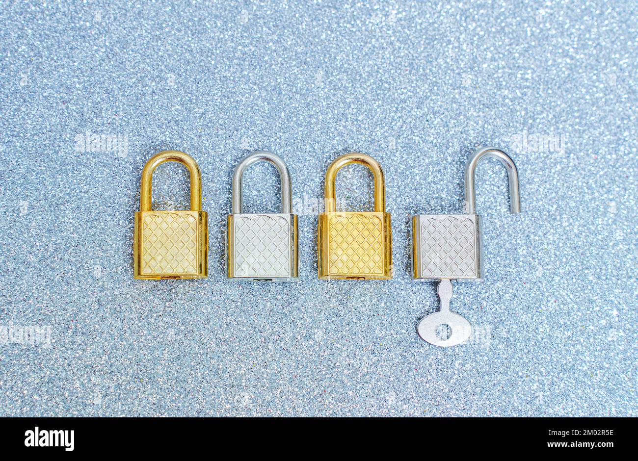 Vier silberfarbene und goldfarbene Vorhängeschlösser auf glitzerndem Hintergrund. Ein Vorhängeschloss wird mit einem Schlüssel geöffnet. Stockfoto