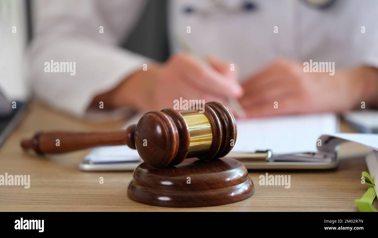 Hölzerner Hammer des Richters auf dem Tisch Nahaufnahme, und verschwommener Arzt, der Papierkram in der medizinischen Klinik erledigt. Stockfoto