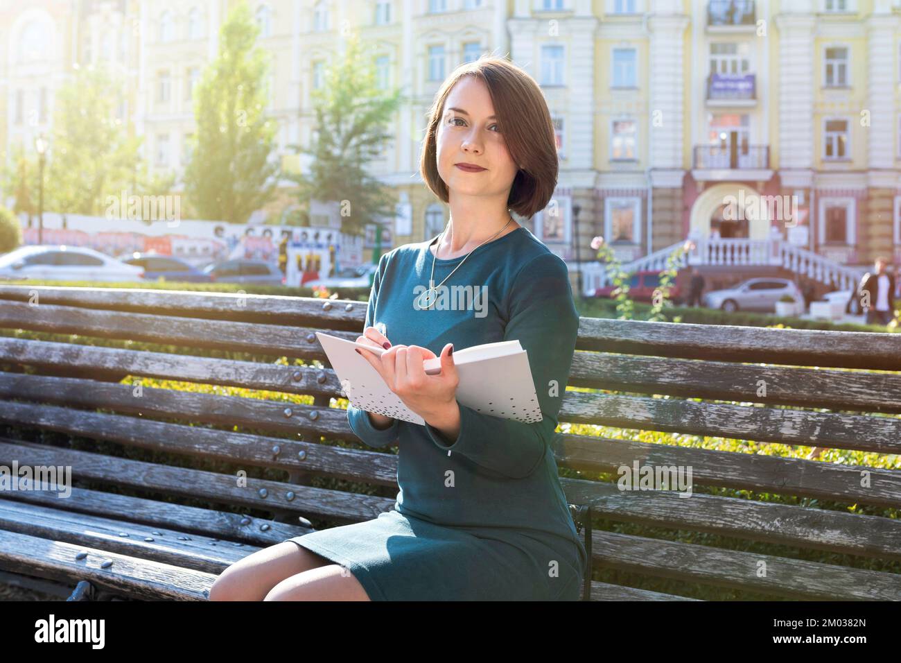 Außenporträt einer Studentin im Freien. Ein junges Mädchen in einem Park, das auf einer Bank sitzt und ein Lehrbuch in der Hand hält Stockfoto