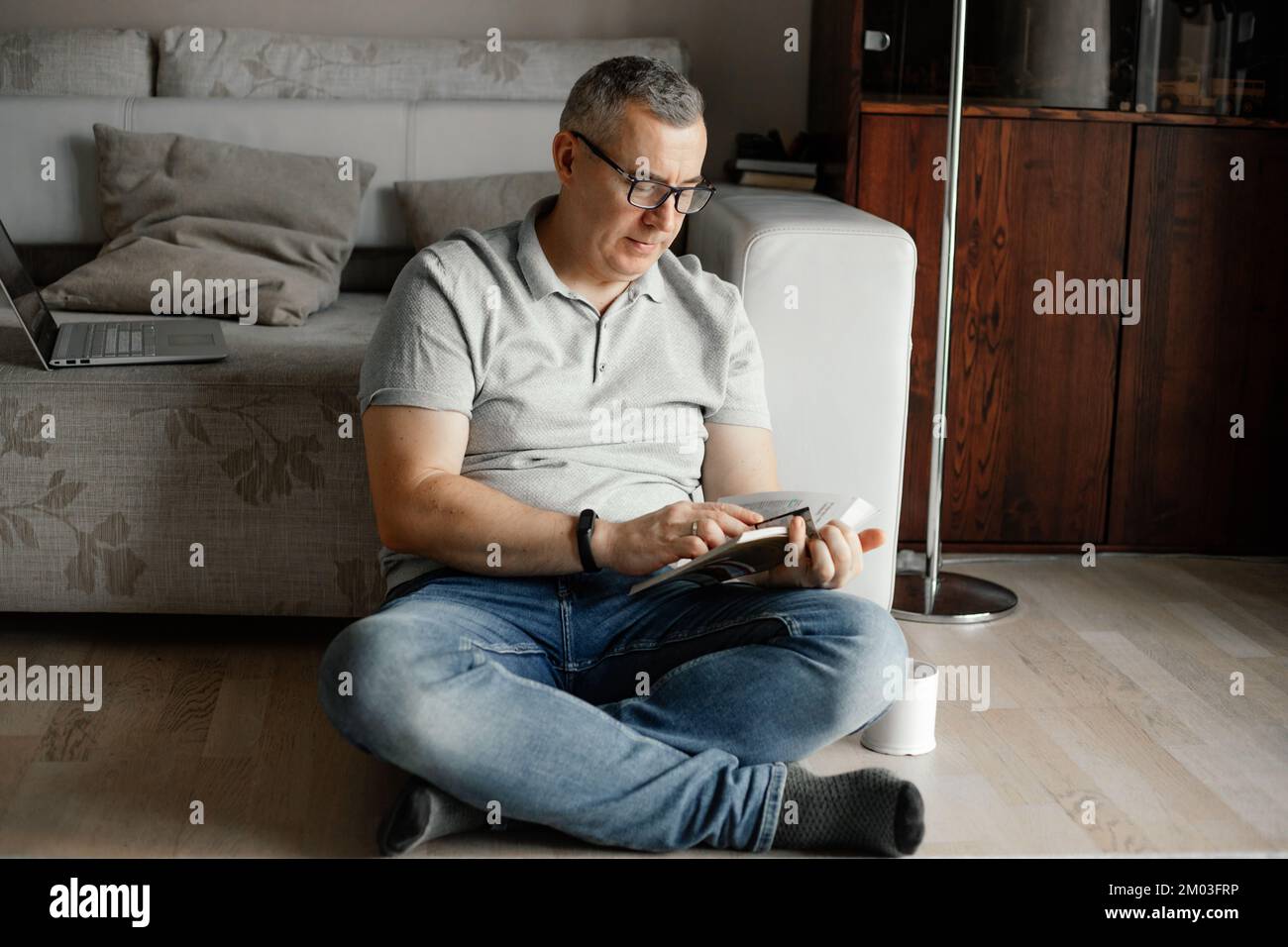 Porträt eines konzentrierten Mannes mittleren Alters, der mit gekreuzten Beinen neben dem Sofa auf dem Boden sitzt und zu Hause ein Buch hält. Stockfoto