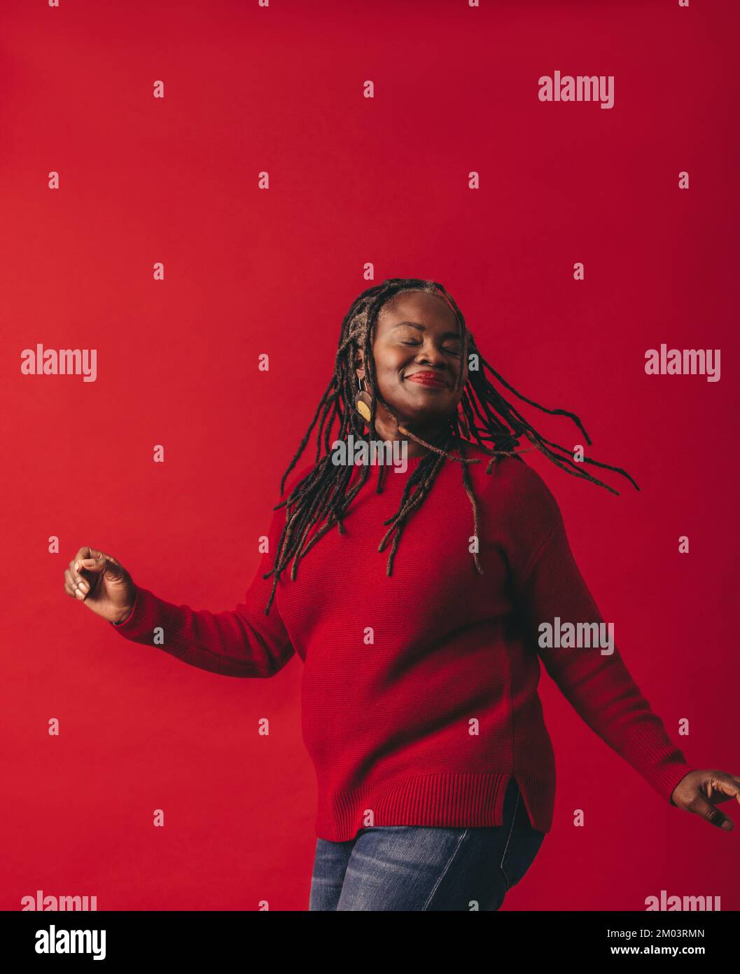 Fröhliche schwarze Frau tanzt und peitscht ihre Dreadlocks, während sie vor einem roten Hintergrund steht. Glückliche reife Frau, die Spaß hat und ihre Natura umarmt Stockfoto
