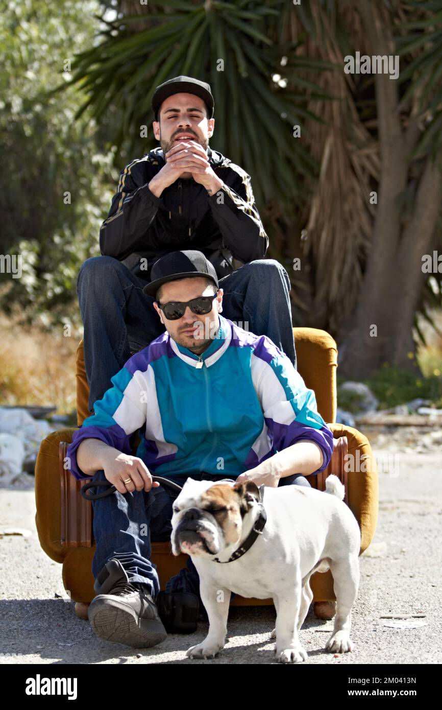 Urbaner Lebensstil. Zwei Gangster sitzen auf einer Couch, die sich in der Mitte einer Straße mit ihrem Hund befindet. Stockfoto