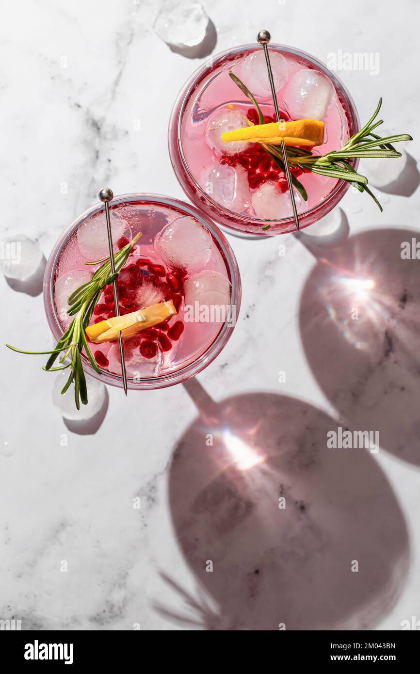 Zwei Weingläser aus Granatapfel, Eiscocktail mit Rosmarin und Zitrone auf weißem Marmorhintergrund, isoliert mit klaren Schatten in rauem Licht Stockfoto