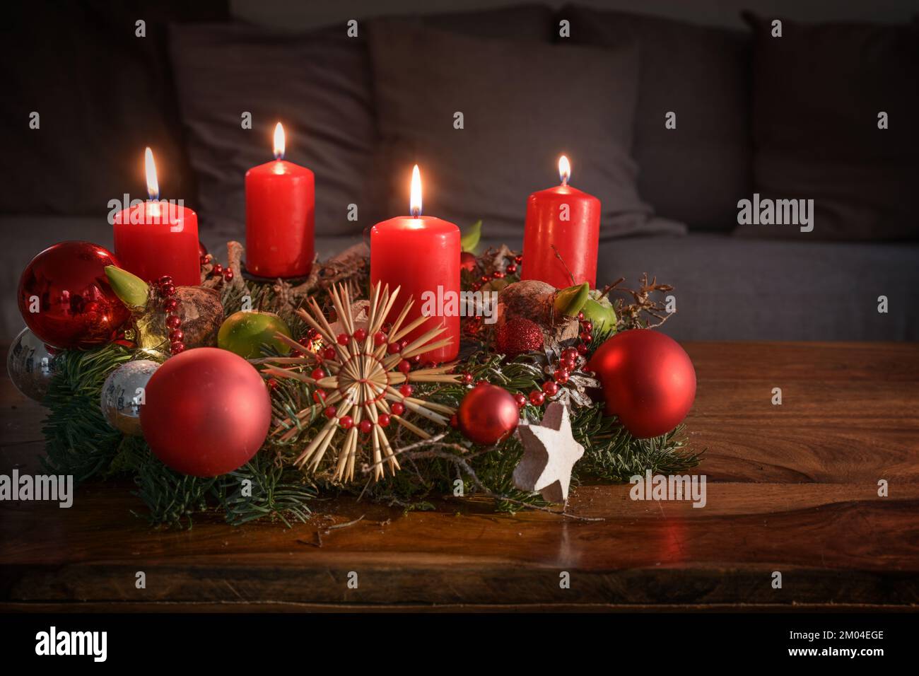 Adventskranz mit vier brennenden roten Kerzen und Weihnachtsdekoration auf einem Holztisch vor der Couch, festliches Heimdekor für den vierten Sonntag Stockfoto