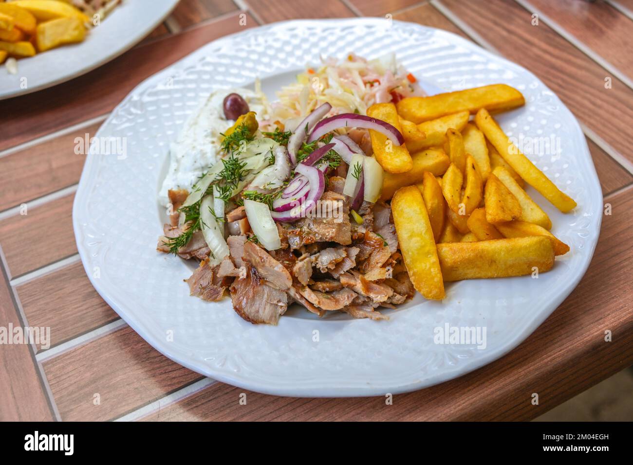 Gyros, griechisches Gericht aus aufgeschnittenem Fleisch, geröstet auf einem drehenden Spieß, serviert mit Pommes Frites, Krautsalat, Tzatziki und Zwiebeln auf einem weißen Teller, Holz im Freien Stockfoto