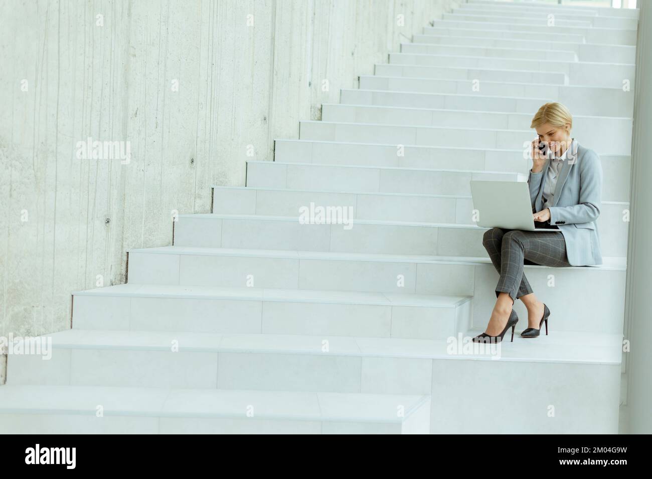 Geschäftsfrau, die auf der modernen Bürotreppe mit einem Mobiltelefon an einem Laptop arbeitet Stockfoto