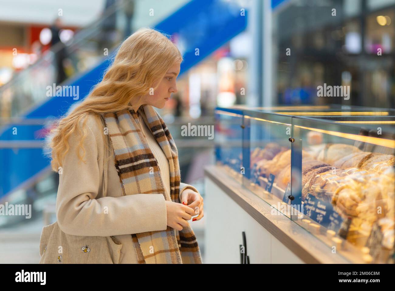 Eine Frau in weißem Mantel und Schal, die auf die Bäckerei mit verschiedenen Kuchenstücken schaut. Konsumerismus, gesunde Lebensmittelkonzepte. Stockfoto
