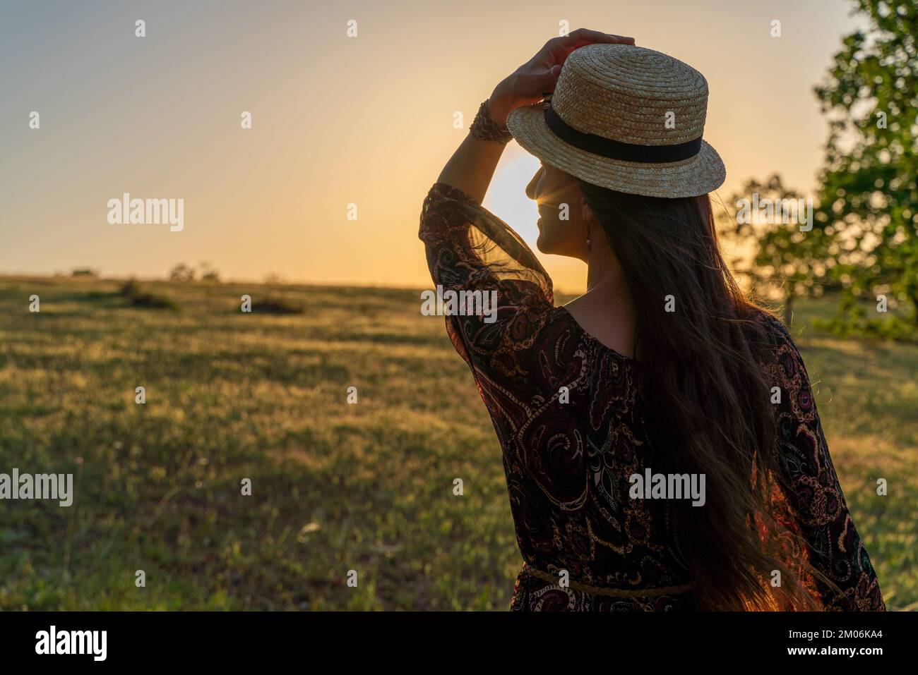 Eine glückliche Frau reist allein im Wald bei Sonnenuntergang, in einem langen Kleid, Hut und einem Blumenstrauß. Frohe Ferien. Rückansicht einer glücklichen Frau Stockfoto